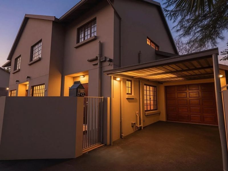 Front elevation of a townhouse featuring a paved driveway, covered carport, secure boundary wall with pedestrian gate, and exterior lighting at dusk.