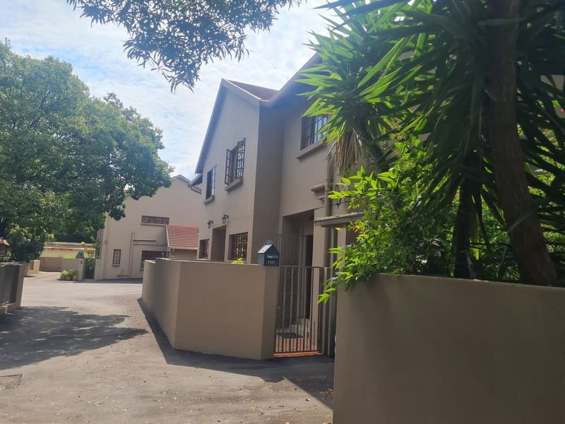 A view of the townhouse exterior featuring a paved driveway, perimeter wall, and secure gated entrance with established surrounding trees.