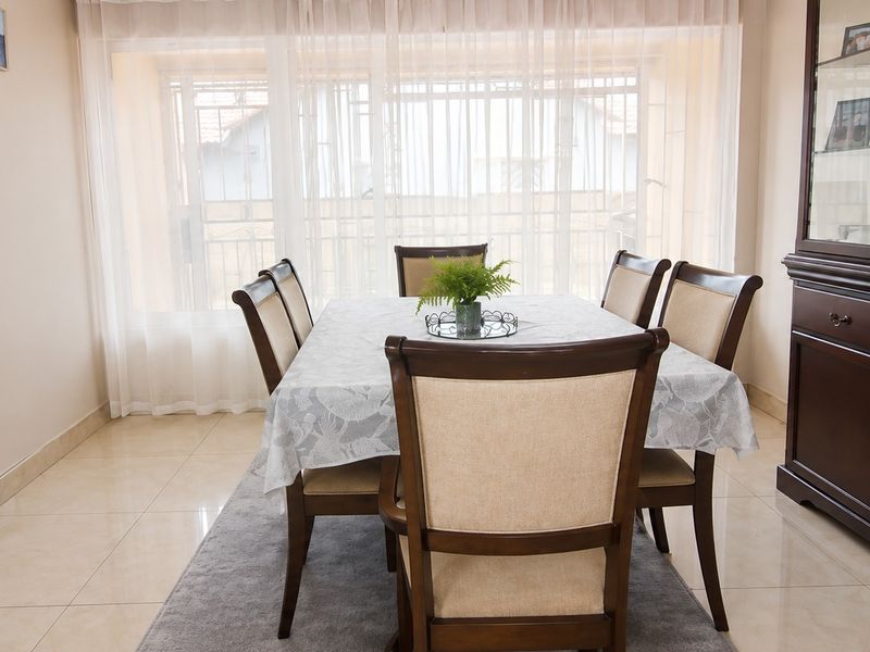 A well-maintained dining room featuring neutral tiled flooring and a large window fitted with security bars, providing a bright and functional space.