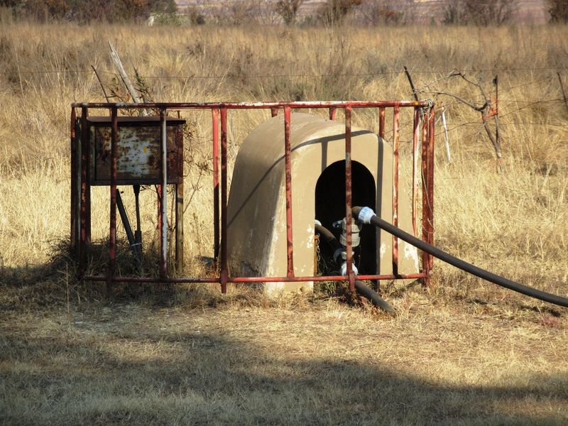 Outdoor borehole pump installation featuring a concrete housing, protective metal fencing, and associated electrical and water piping infrastructure.