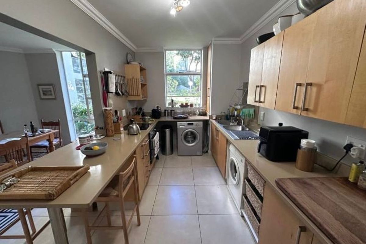 A functional galley-style kitchen featuring light wood cabinetry, laminate countertops, tiled flooring, and an integrated washing machine with an adjoining breakfast bar area.