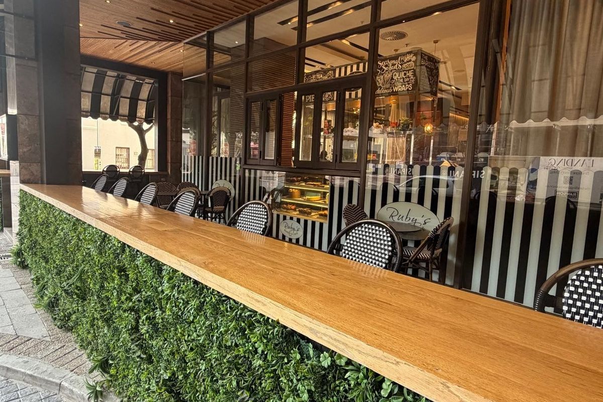 A modern retail storefront featuring a wooden bar counter with integrated artificial greenery, large glass windows, and a covered outdoor seating area.