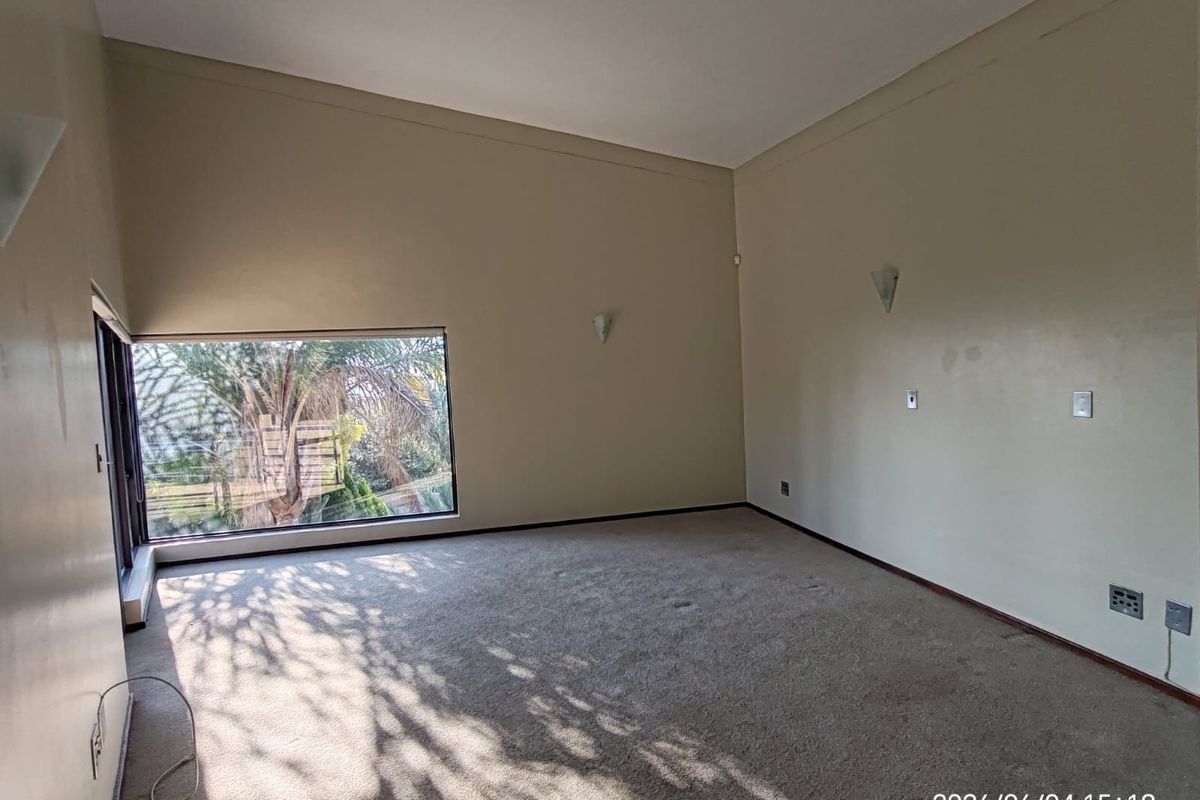 A spacious, empty living room featuring high ceilings, neutral wall tones, carpeted flooring, and a large window providing natural light and garden views.