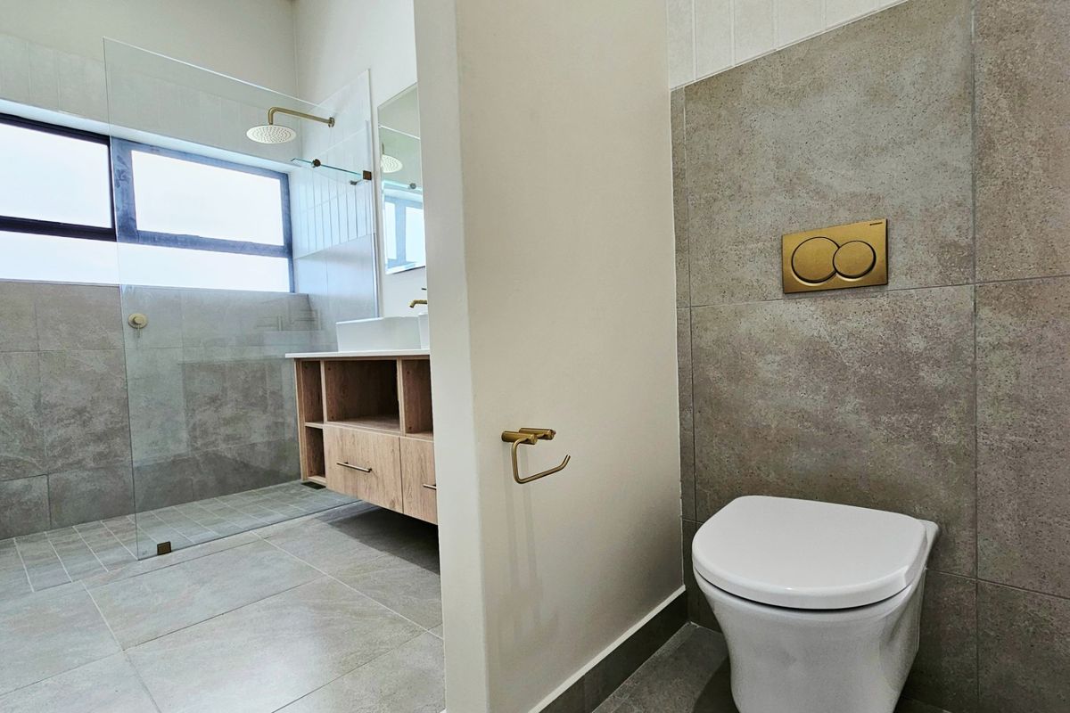 Modern bathroom featuring a wall-mounted toilet with gold flush plate, walk-in glass shower, and a floating wooden vanity with basin.