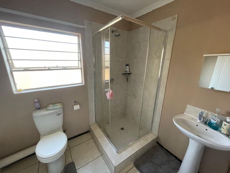 A functional bathroom featuring a corner glass shower enclosure, pedestal basin, and toilet, with tiled flooring and a window for natural light.