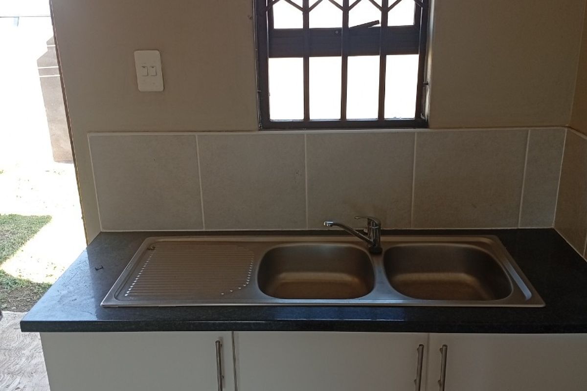 A functional kitchen sink area featuring a double-basin stainless steel sink, dark countertop, and white cabinetry, positioned beneath a window with security bars.