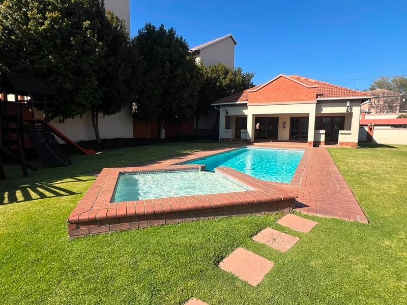 A well-maintained communal pool area featuring a swimming pool, raised spa, brick paving, and a clubhouse building set within a landscaped garden.