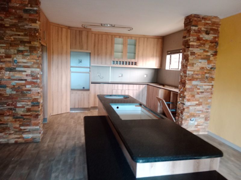 A kitchen space featuring stone-clad pillars, wood-look cabinetry, and a central island with a dark countertop, currently undergoing renovation or awaiting final fixture installation.