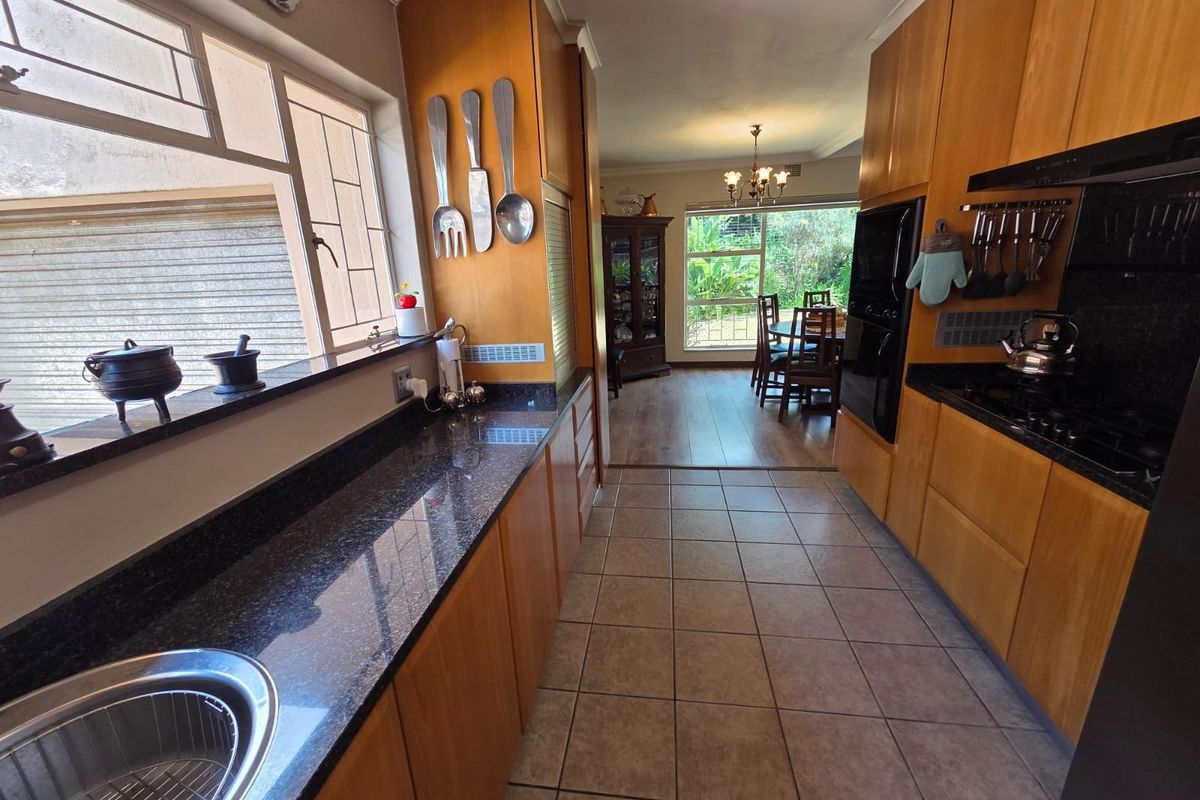 A functional galley-style kitchen featuring wooden cabinetry, dark granite countertops, and tiled flooring, leading into an adjacent dining area with garden views.