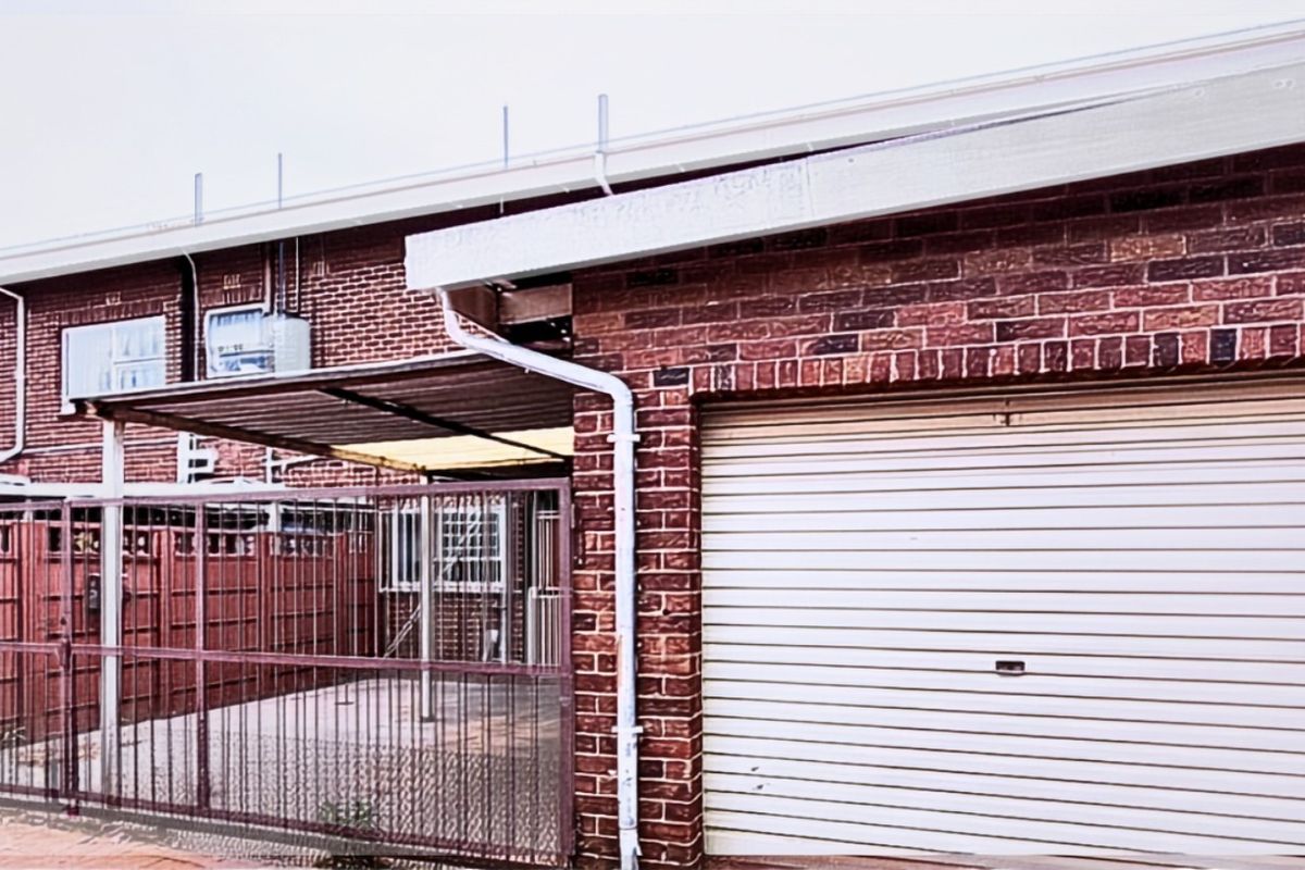 Exterior view of the apartment featuring a brick facade, a secure metal gate, a covered patio area, and a roll-up garage door.
