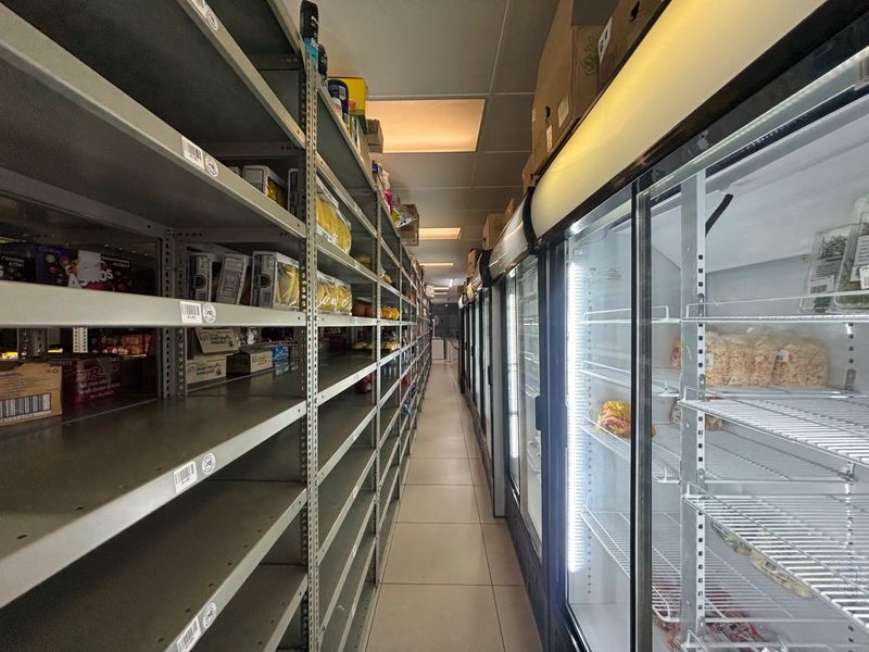 A retail utility aisle featuring rows of metal shelving and commercial glass-door refrigeration units with integrated lighting.