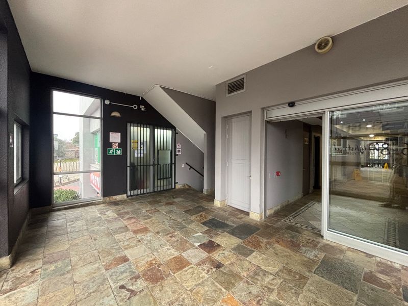 A clean, tiled entrance lobby area featuring a sliding glass door, security gate, and access to a stairwell in a commercial retail building.