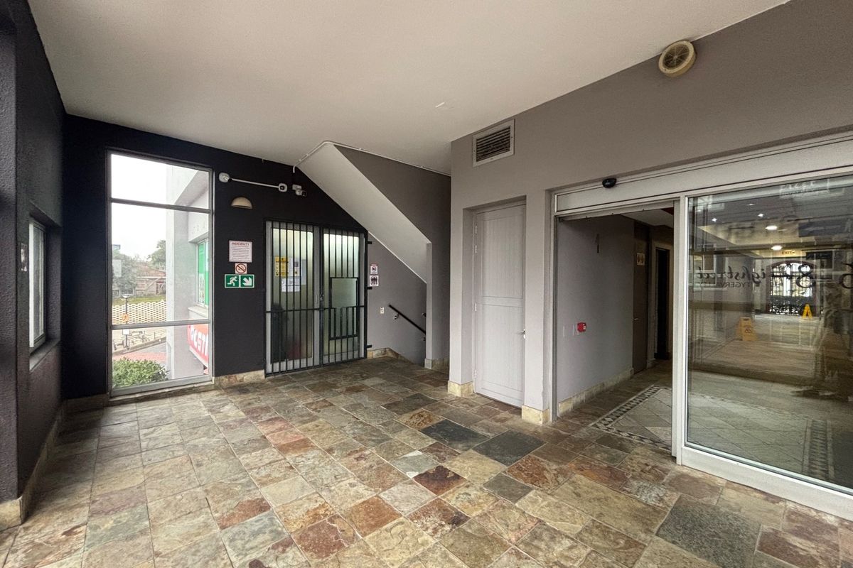 A clean, tiled entrance lobby area featuring a sliding glass door, security gate, and access to a stairwell in a commercial retail building.