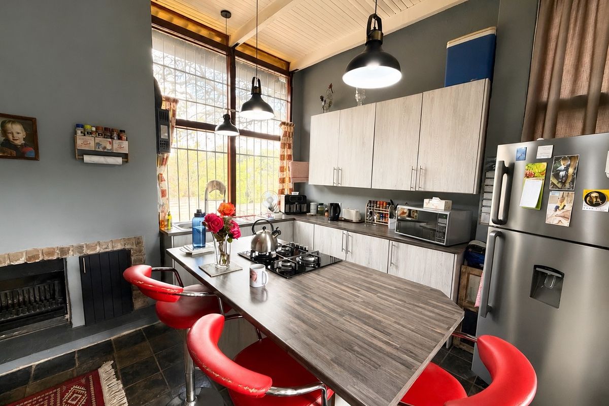 A functional kitchen featuring a central island with a built-in gas hob, light-toned cabinetry, and a cozy fireplace, illuminated by large windows.