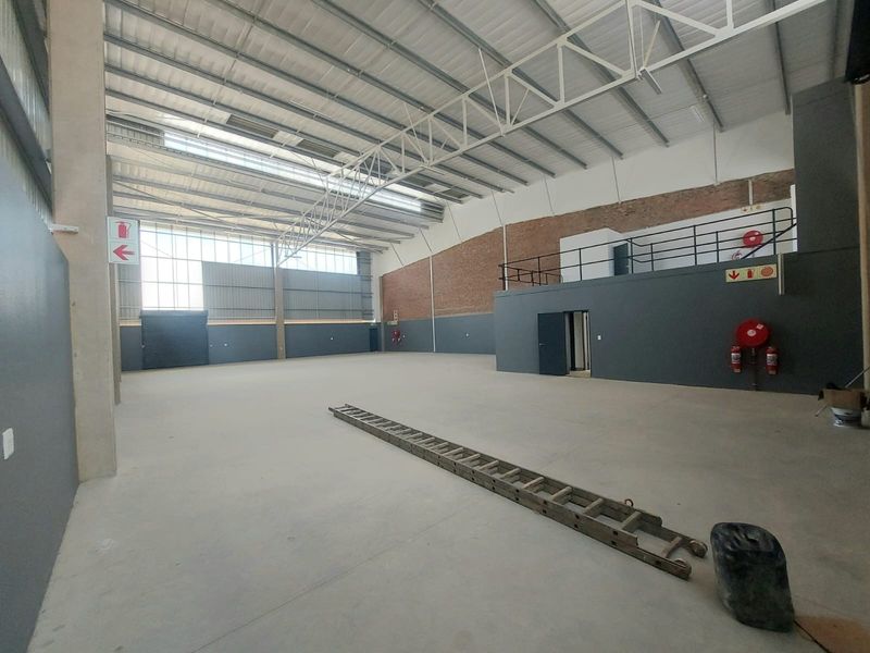 Spacious industrial warehouse unit featuring high ceilings with exposed steel trusses, a mezzanine level, concrete flooring, and a large roller shutter door.