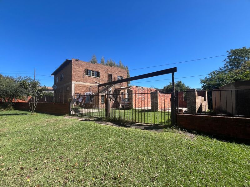 Exterior view of a two-story brick house featuring a large lawn, metal perimeter fencing, and unfinished structural brickwork elements in the yard.