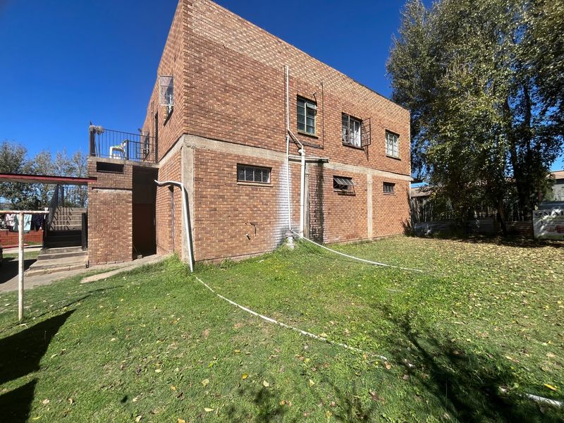 Rear view of a two-story brick house featuring a small balcony, exposed exterior plumbing, and a large backyard area.