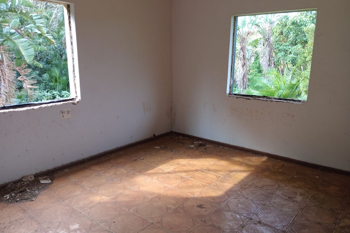 An empty bedroom featuring tiled flooring and two window openings with views of the surrounding garden, requiring significant renovation and repair.
