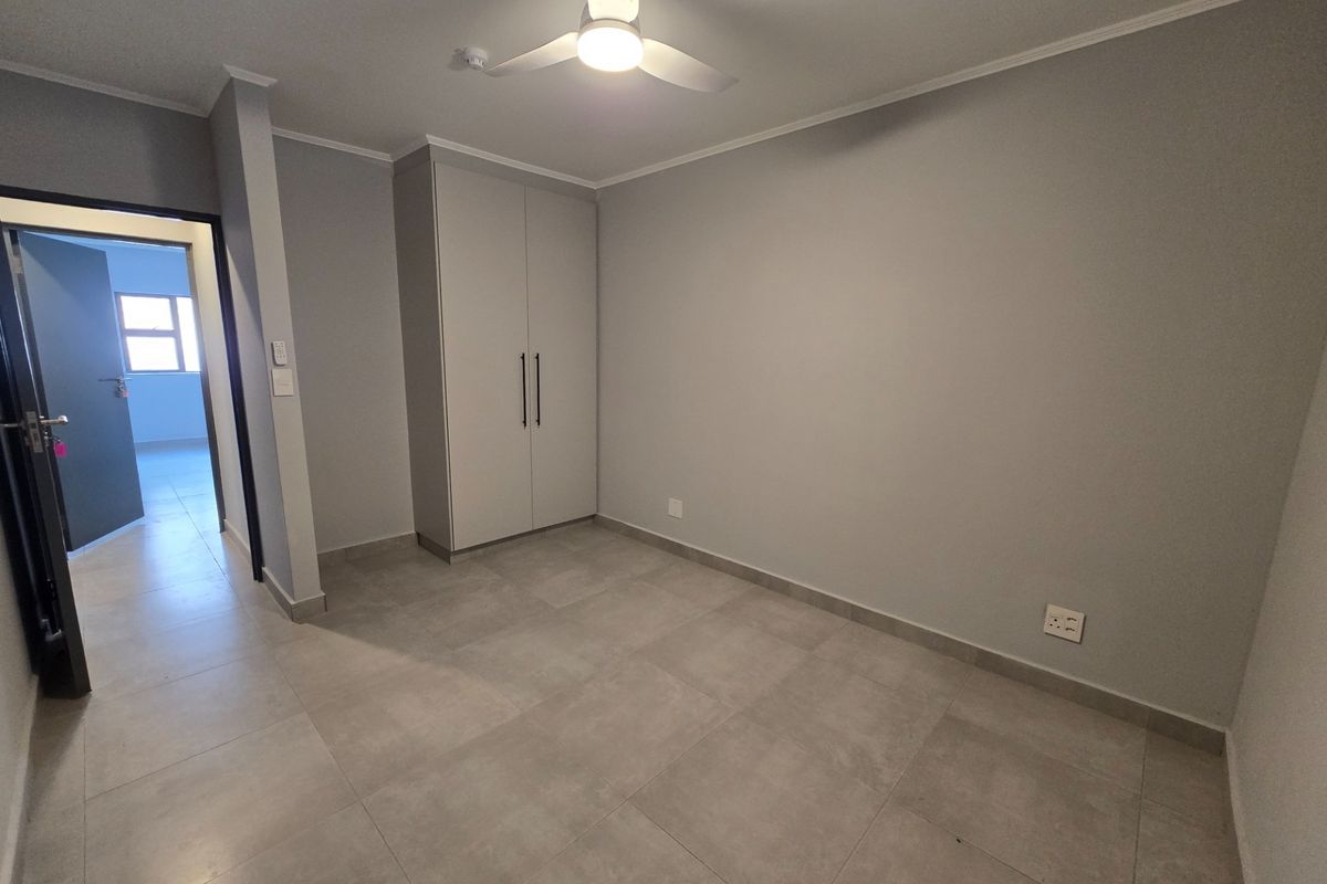 A clean, empty bedroom featuring neutral grey walls, large format floor tiles, a built-in wardrobe, and a modern ceiling fan.