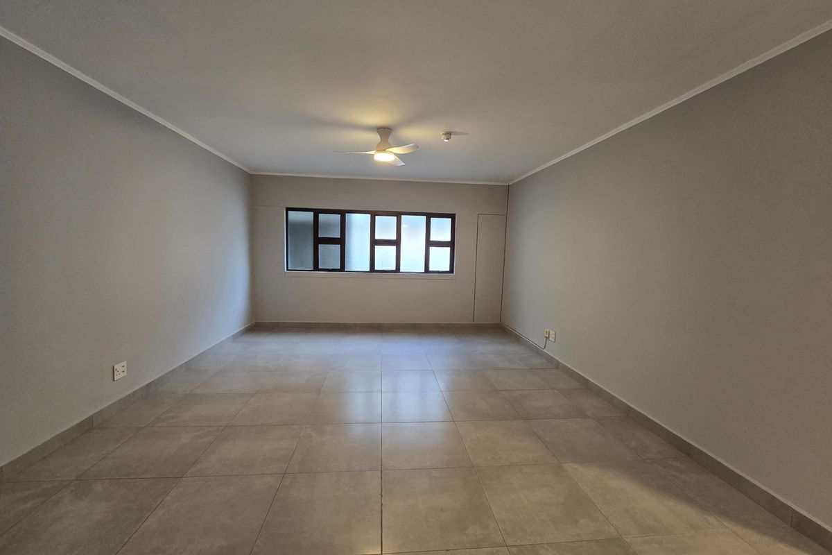 A spacious, empty living area featuring neutral tiled flooring, a ceiling fan, and a large window providing natural light.