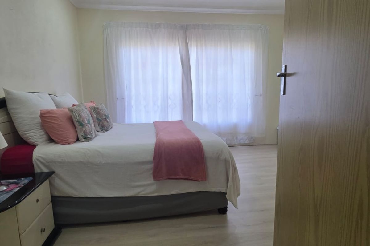 A bedroom featuring light-toned laminate flooring, neutral wall finishes, and a large window dressed with sheer curtains.