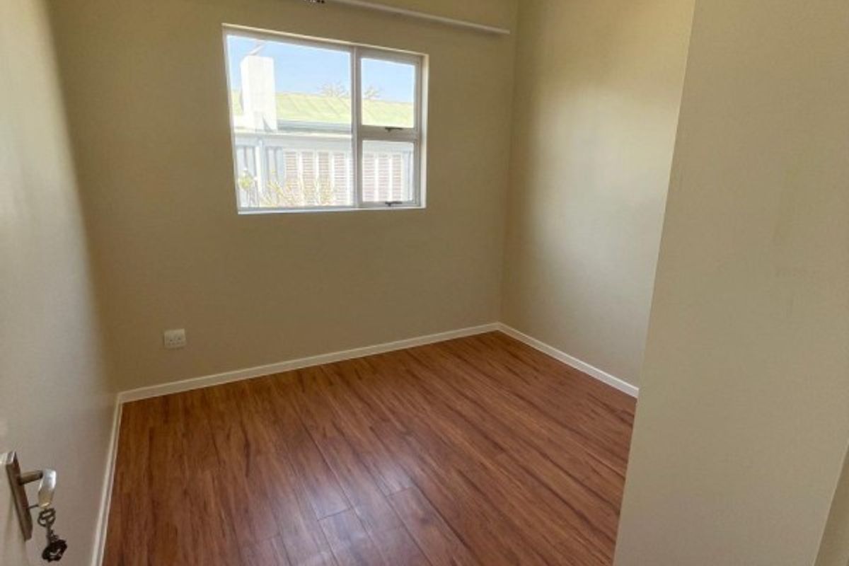 A bright, empty bedroom featuring wood-look laminate flooring, recessed ceiling lighting, and a window with a curtain rail.