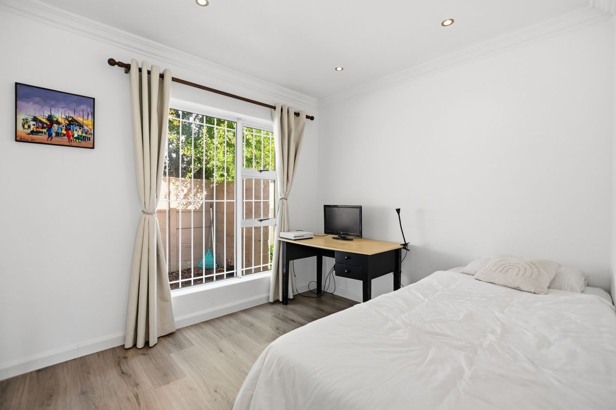 A bright bedroom featuring modern laminate flooring, recessed ceiling lighting, and a window fitted with security bars.