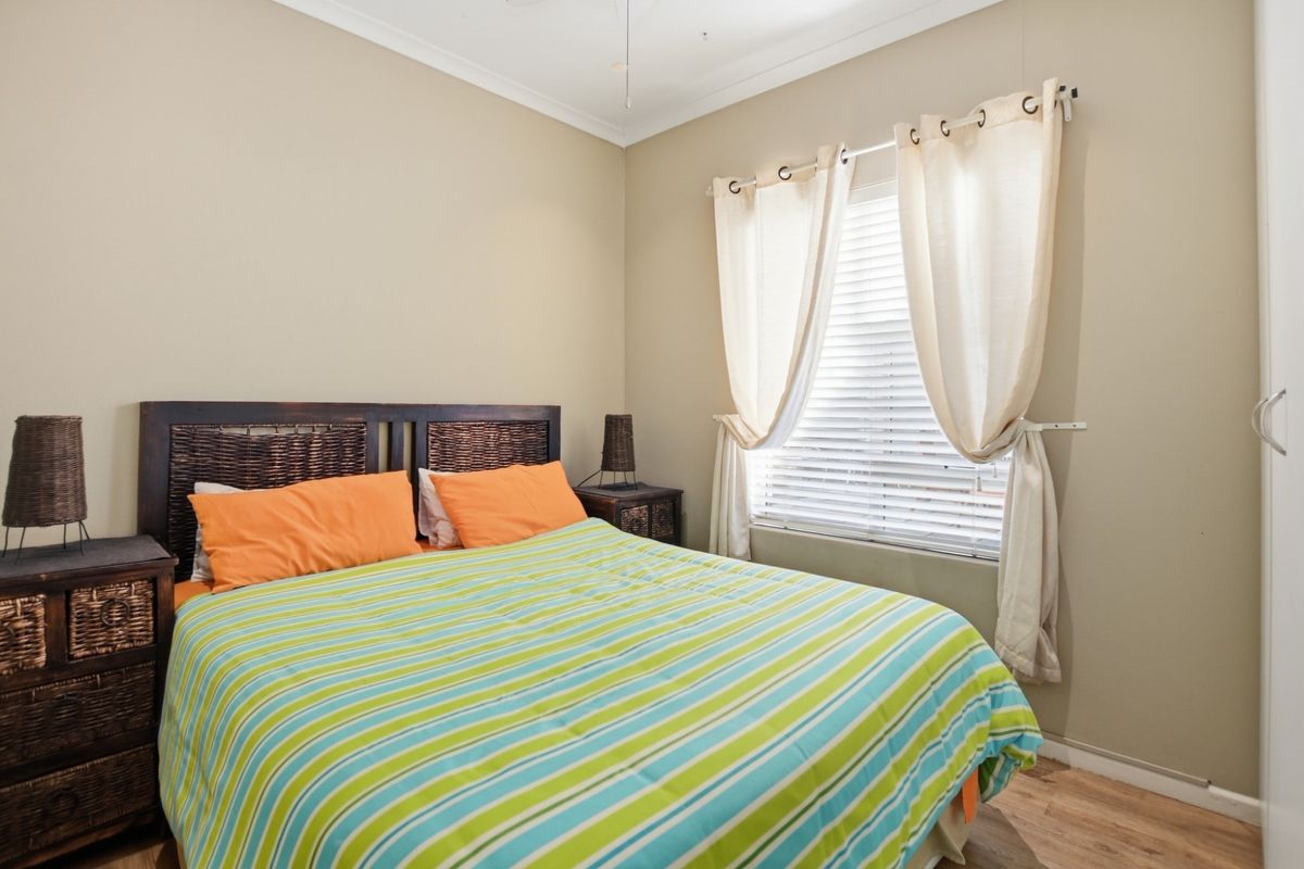 A well-lit bedroom featuring neutral wall tones, wood-look flooring, and a window fitted with venetian blinds and curtain rods.