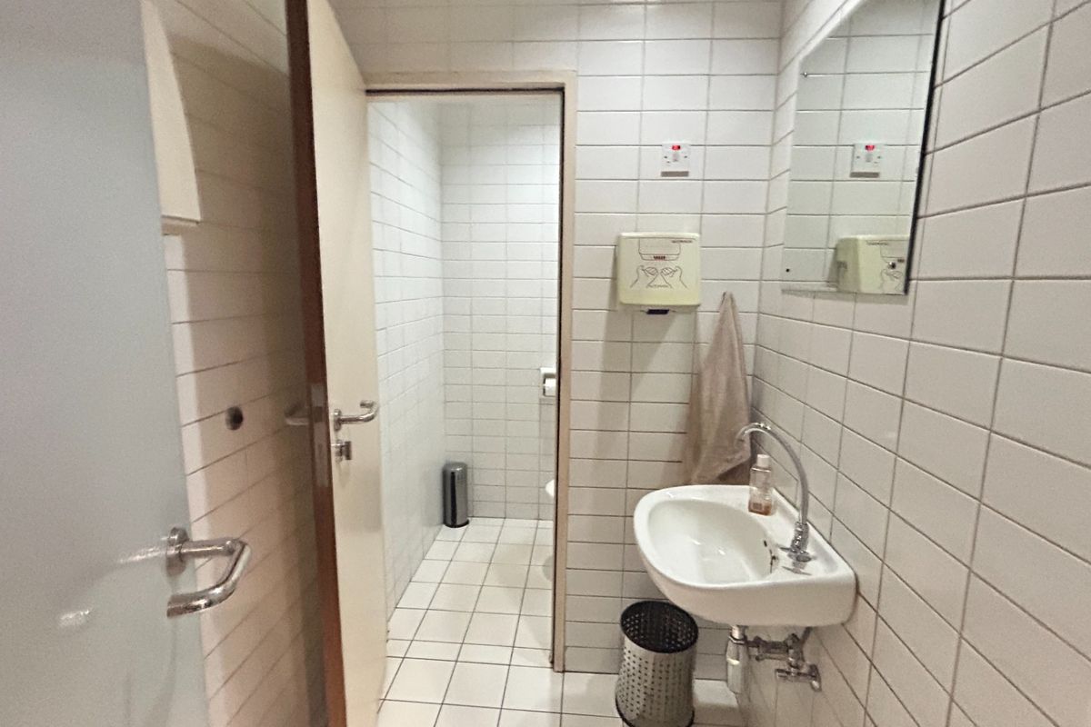 A functional commercial restroom featuring white tiled walls and flooring, a wall-mounted basin, and a separate toilet cubicle.