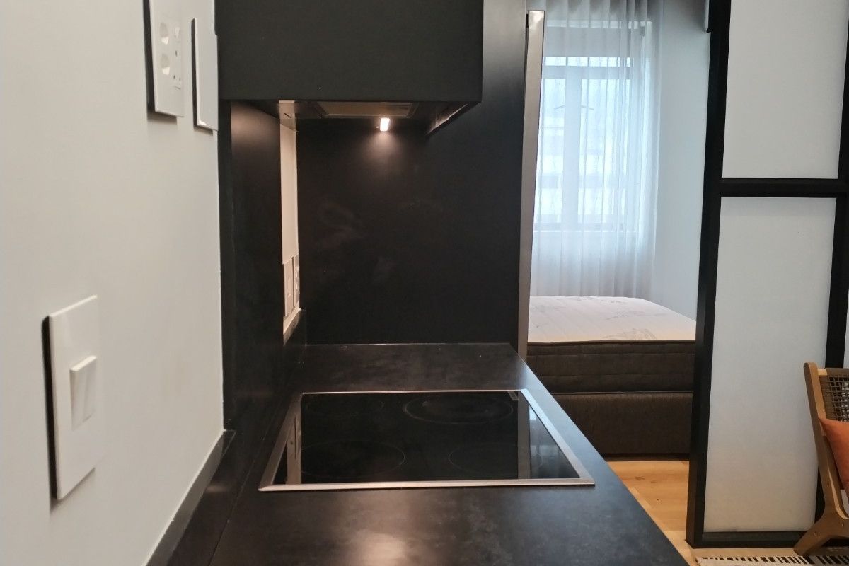 Modern compact kitchen featuring a sleek black countertop, integrated induction cooktop, and built-in extractor fan, separated from the bedroom by a stylish glass partition.