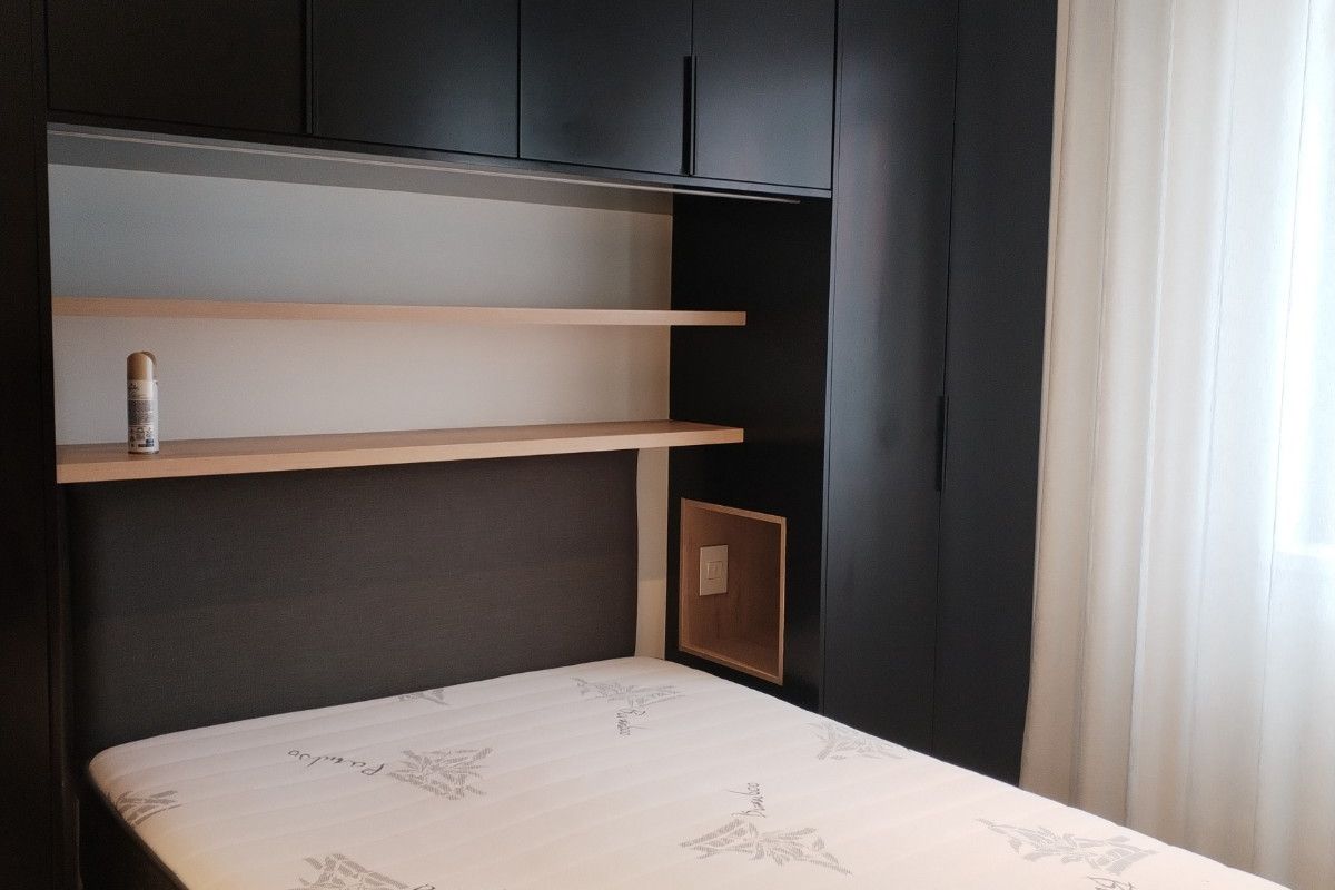 A bedroom featuring modern dark cabinetry with integrated overhead storage, open shelving, and a built-in bedside niche with electrical access.