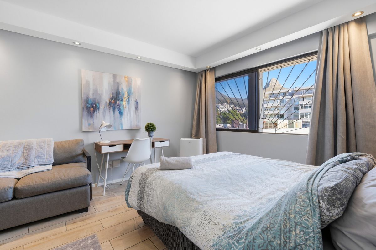 A well-maintained bedroom featuring wood-look tile flooring, recessed ceiling lighting, and a large window with security bars offering an urban view.