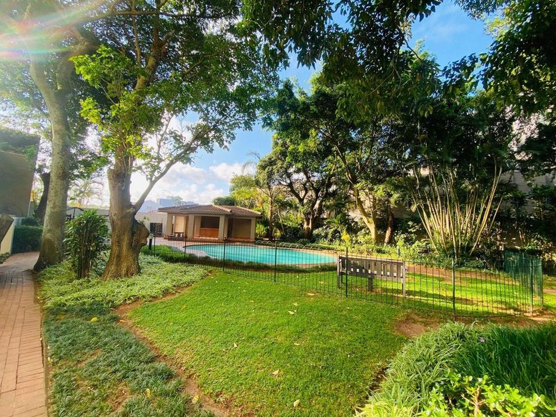 A well-maintained communal pool area featuring a swimming pool, poolside pavilion, and lush landscaped gardens with mature trees.