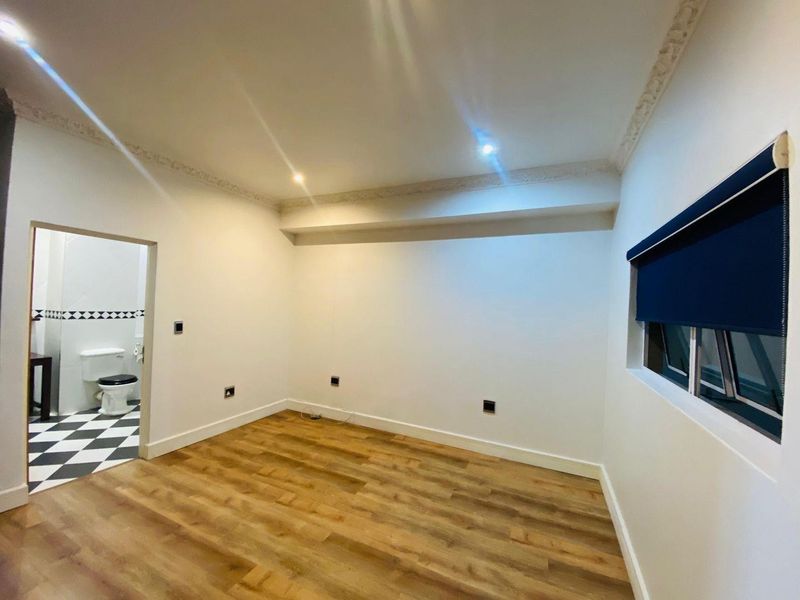 A bright, empty bedroom featuring laminate flooring, decorative ceiling cornice, and a roller blind, with an adjoining bathroom visible through the doorway.