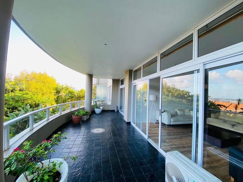 Spacious covered balcony featuring dark tiled flooring, glass balustrades, and large sliding glass doors providing seamless indoor-outdoor flow.