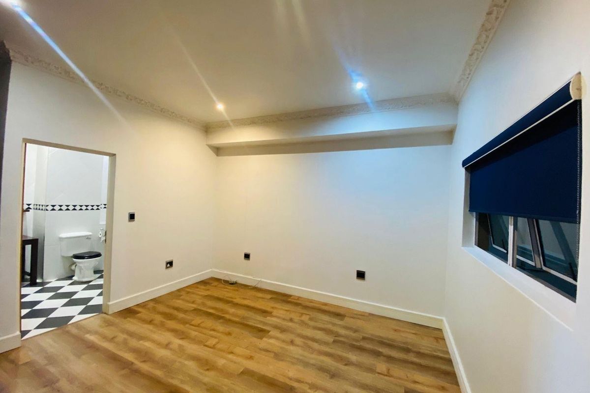 A bright, empty bedroom featuring laminate flooring, decorative ceiling cornice, and a roller blind, with an adjoining bathroom visible through the doorway.