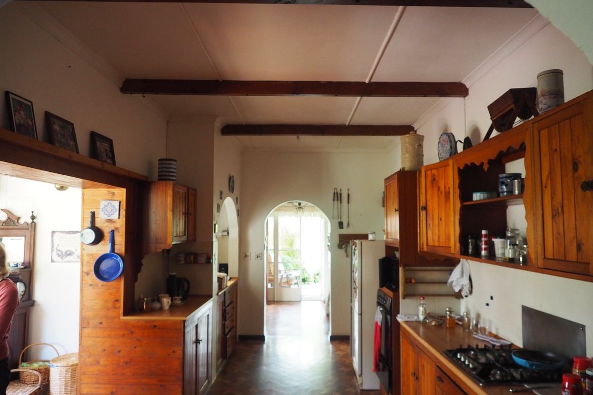 A rustic farmhouse kitchen featuring timber cabinetry, exposed ceiling beams, and a gas hob, leading through an arched doorway to an adjacent living space.