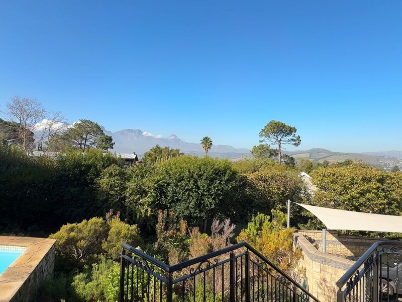 Elevated outdoor view featuring a swimming pool, stone retaining walls, and a shade sail, overlooking lush greenery and distant mountain ranges.