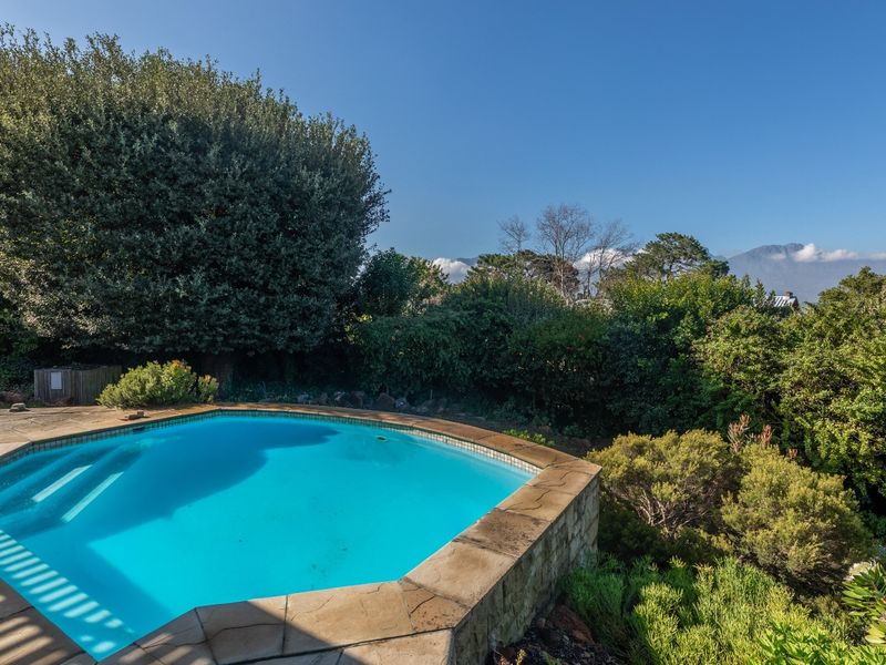 A well-maintained swimming pool area featuring stone coping and a paved deck, surrounded by lush garden landscaping and mountain views.