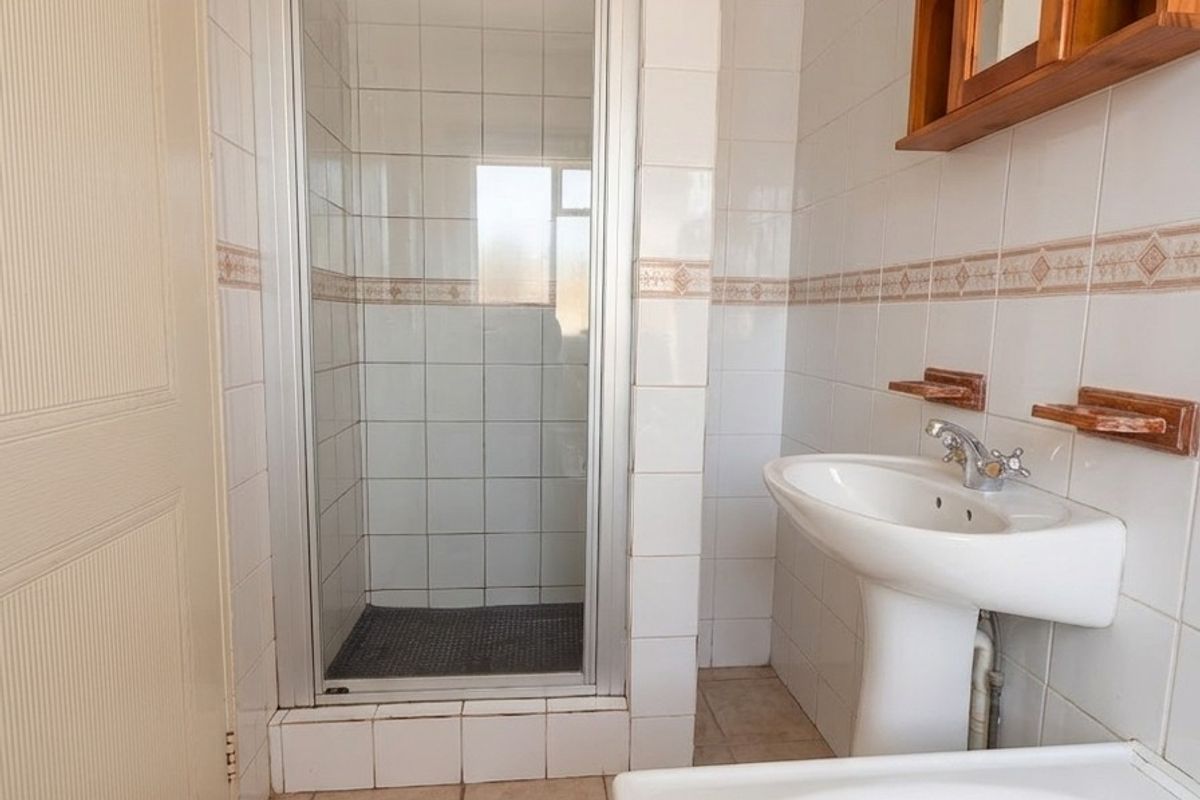 A functional bathroom featuring a pedestal basin, a shower enclosure, and neutral wall and floor tiling, offering a clean and practical layout.