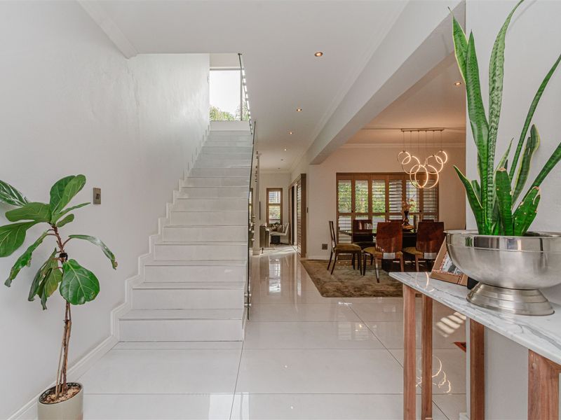 Bright and modern entrance hall featuring polished tiled flooring, a sleek staircase with glass balustrade, and recessed lighting leading into the dining area.