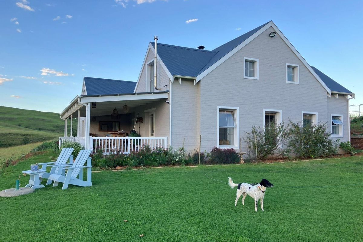 A charming double-story house featuring a covered patio, metal roof, and well-maintained garden, set against a scenic rural landscape.