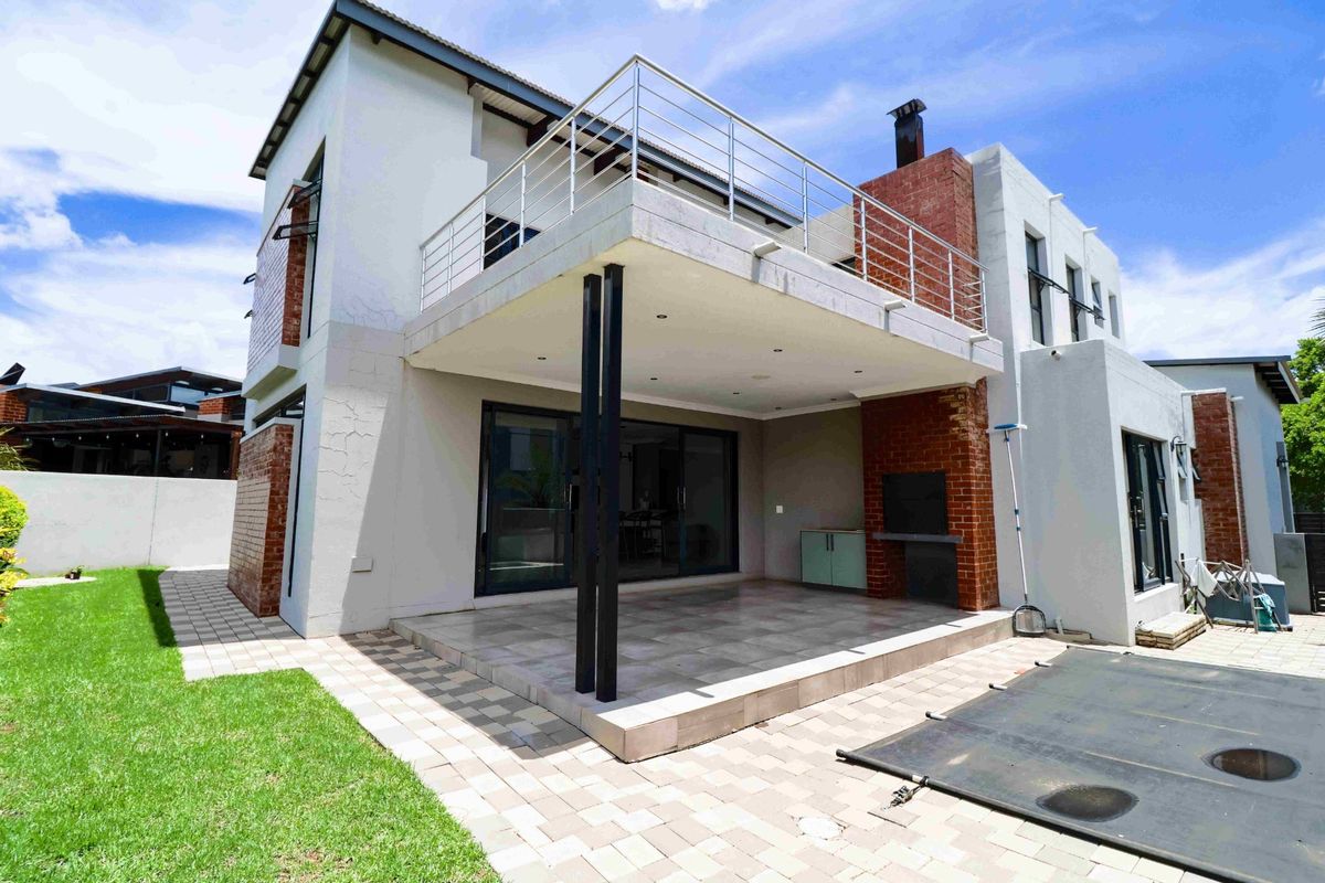 Rear view of a modern two-story house featuring a covered patio with a built-in braai, upper balcony with metal railings, and paved garden area.