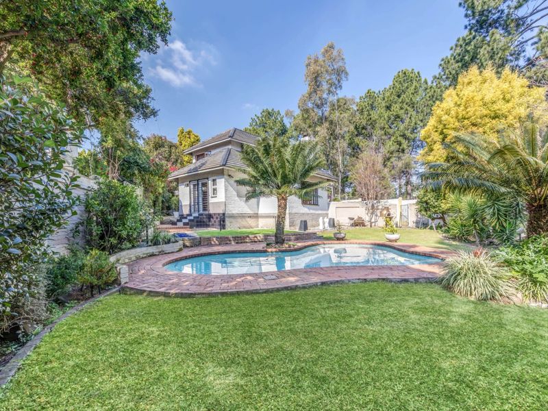 A well-maintained rear garden featuring a swimming pool with brick paving, lush lawn, and mature landscaping surrounding the house.