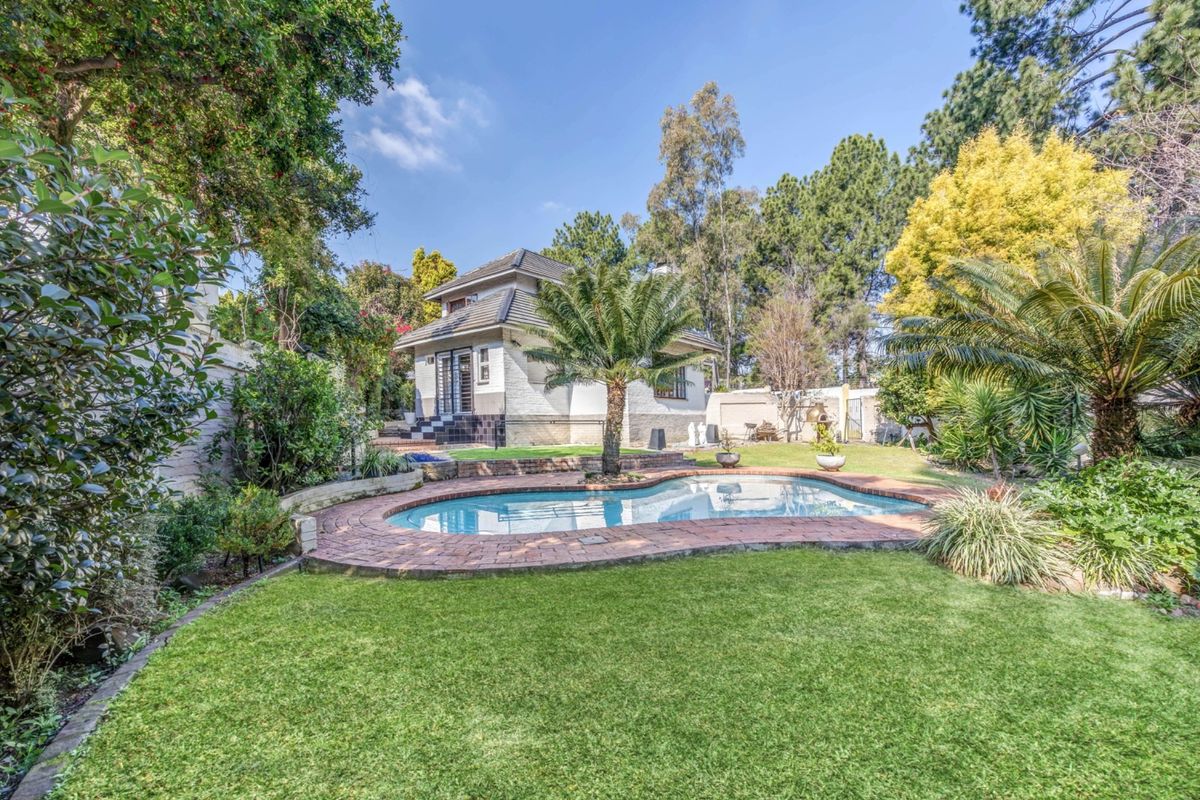 A well-maintained rear garden featuring a swimming pool with brick paving, lush lawn, and mature landscaping surrounding the house.
