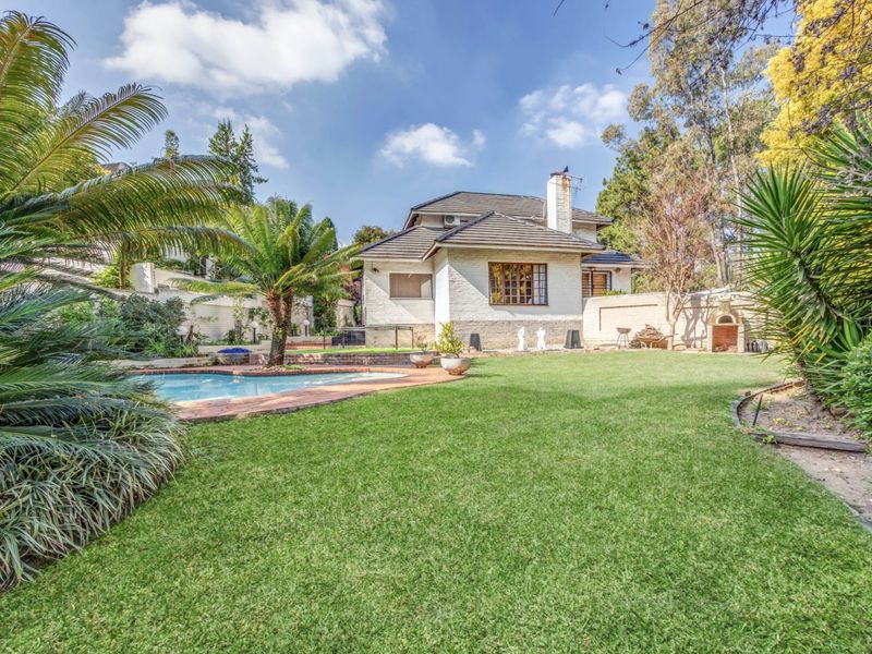 Spacious rear garden featuring a swimming pool, established landscaping, and a brick-built outdoor fireplace, set against the rear elevation of the house.