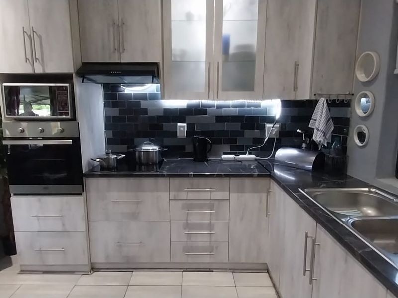 Modern kitchen featuring light wood-grain cabinetry, dark stone-look countertops, a black subway tile backsplash, built-in oven, and a double stainless steel sink.