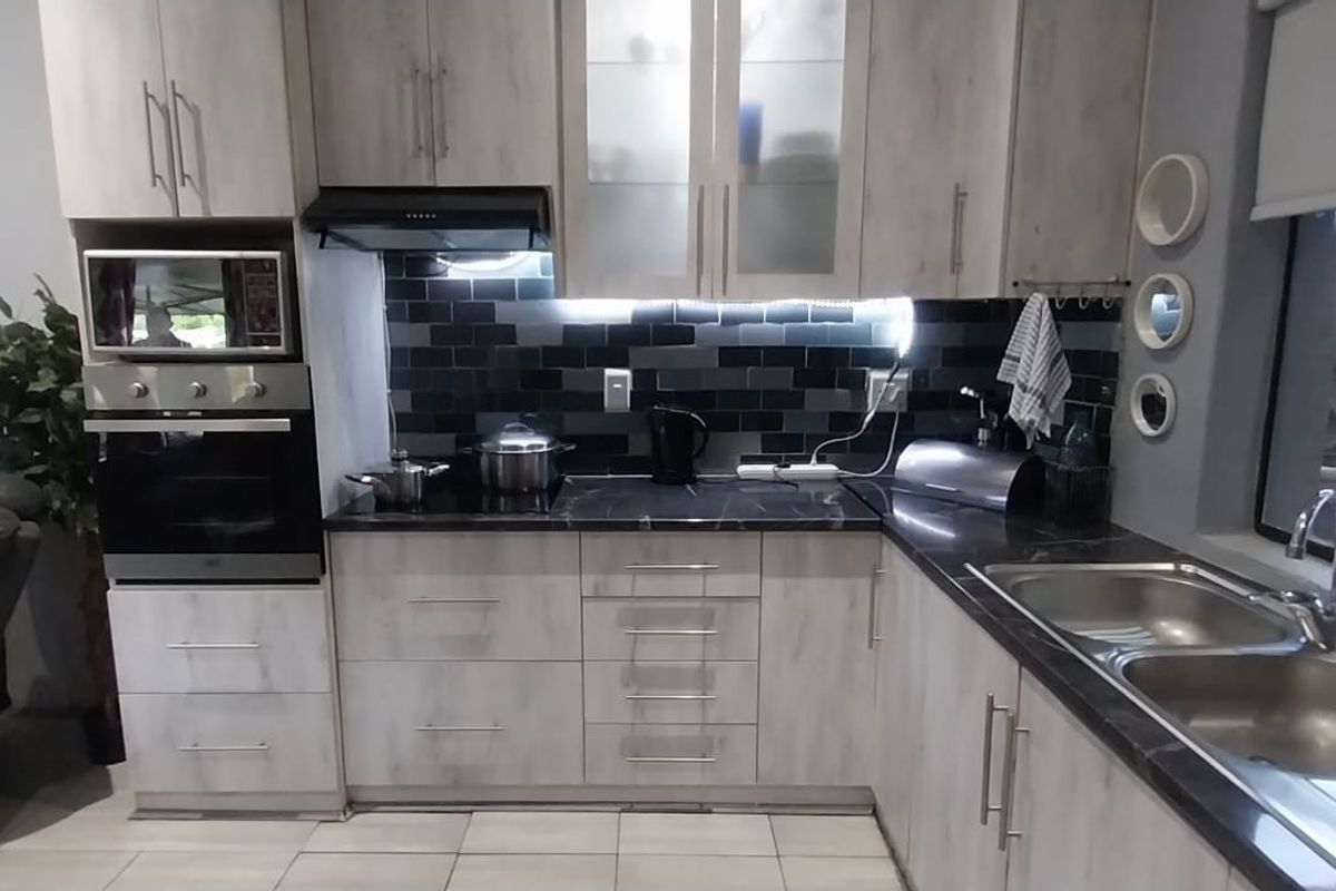 Modern kitchen featuring light wood-grain cabinetry, dark stone-look countertops, a black subway tile backsplash, built-in oven, and a double stainless steel sink.