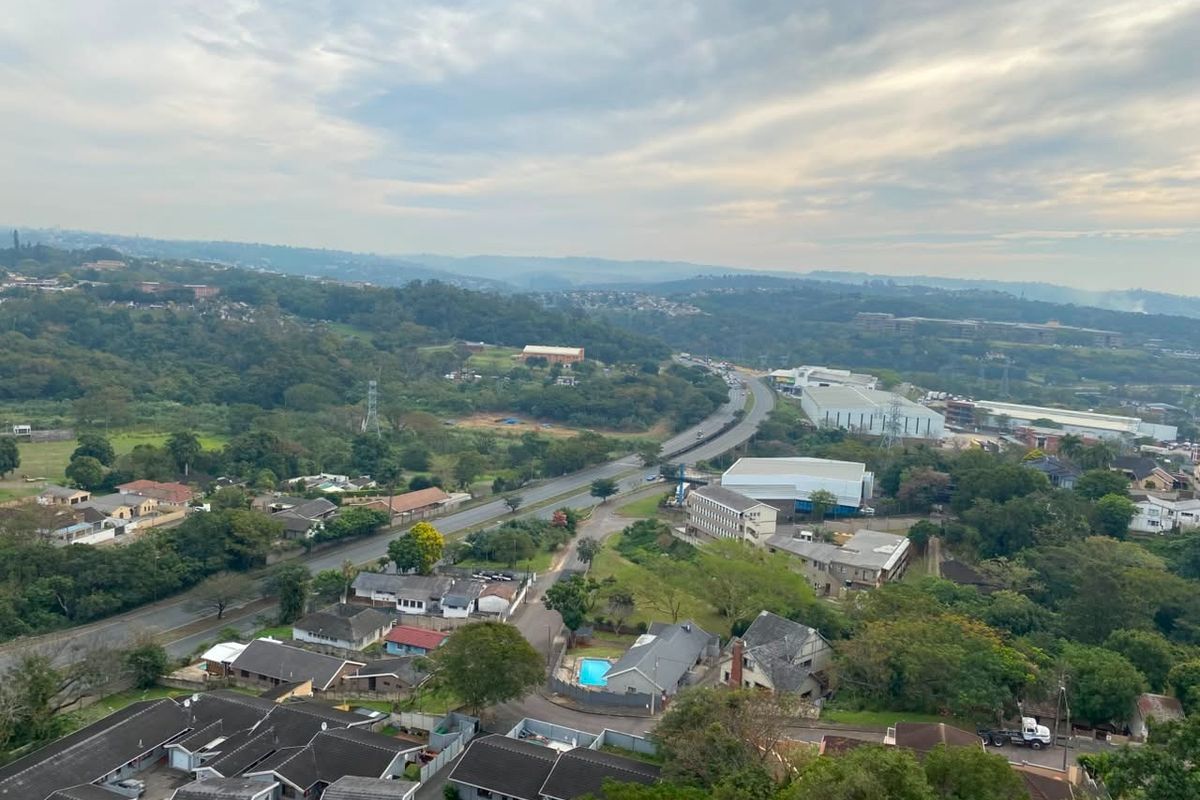 An elevated aerial view overlooking a suburban landscape, featuring residential housing, green spaces, and a main road winding through the area.
