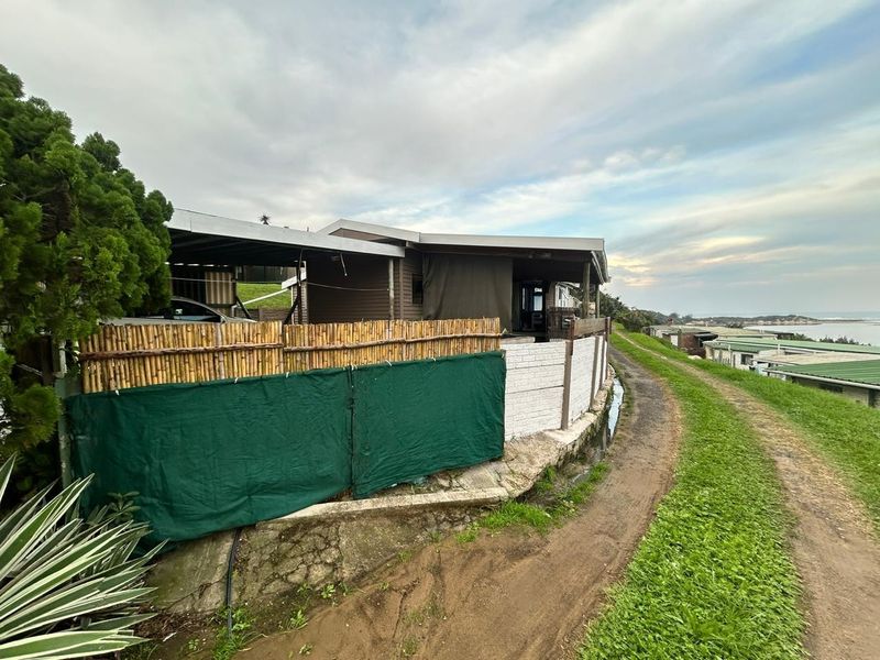 Exterior view of a coastal property featuring a covered patio area, bamboo screening, and a concrete retaining wall along a dirt access path.